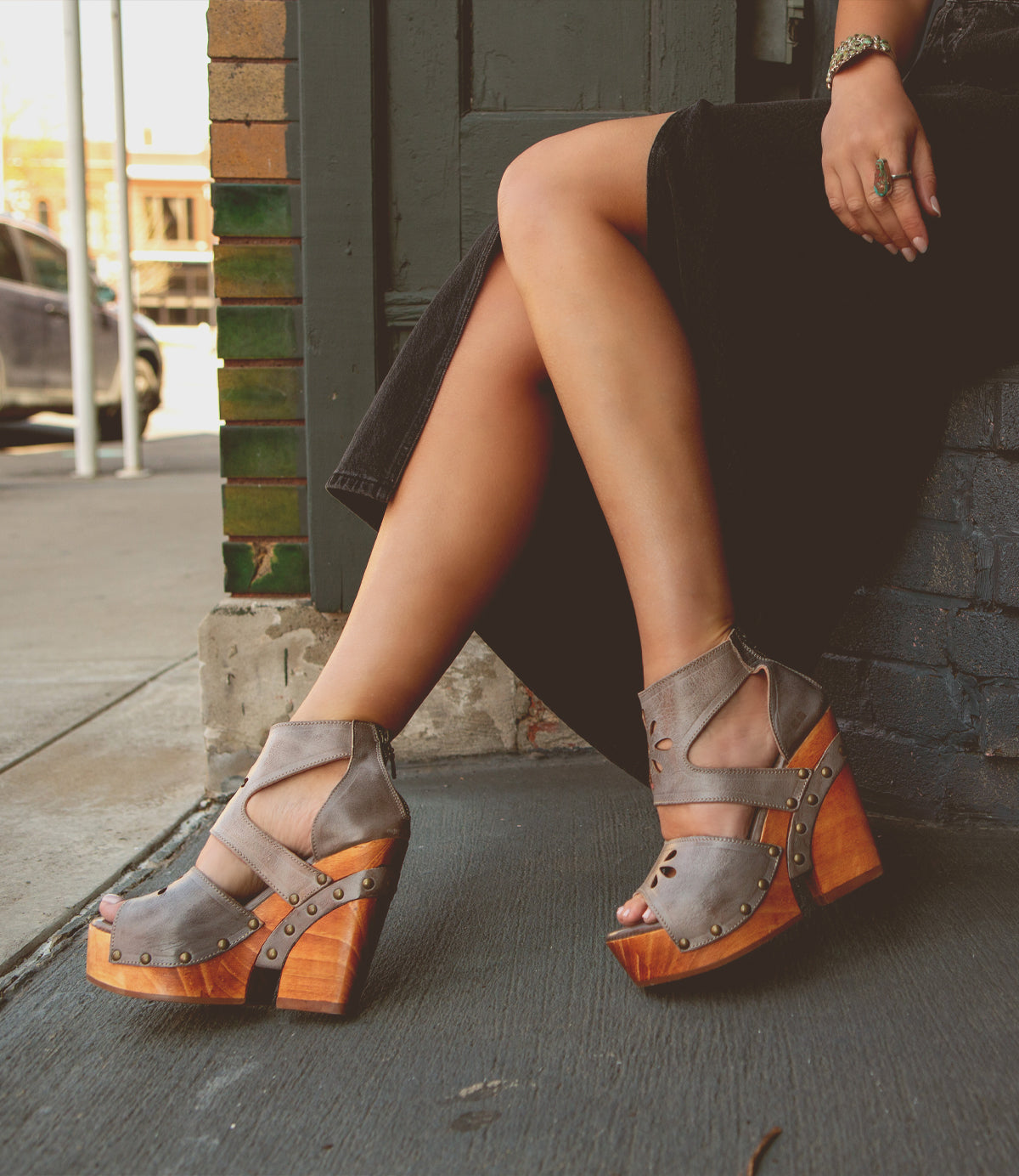 A person sitting on a sidewalk with their legs crossed, wearing Lucrative studded, wooden wedges from Bed Stu with zipper closure.