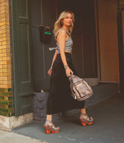 A woman walking on a sidewalk carrying a Bed Stu Lucrative leather shoulder bag with a zipper closure.