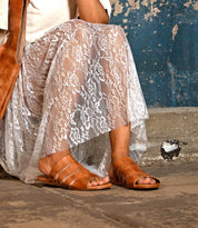A person wearing a white lace skirt and Bed Stu's Margin tan leather upper slide sandals sits on a concrete step next to a worn blue wall, making for an effortless summer look.