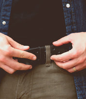 A person is fastening a Bed Stu Mccoy vegetable-tanned leather belt through the belt loops of their beige pants. They are wearing a denim shirt with metal buttons over a black shirt, showcasing a timeless design with modern aesthetics.
