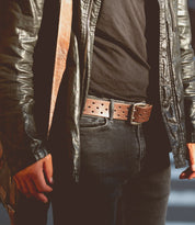 Close-up of a person exuding casual cool style, wearing a black leather jacket, black shirt, dark jeans, and the Proem brown perforated belt with a vintage metal buckle from Bed Stu, standing with arms at their sides.