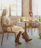 Two people sit at a round table with food and drinks by a large window in a sunlit room. Both are dressed in light-colored, casual clothing. One of them wears the striking Meadow handcrafted leather boots by Bed Stu, adorned with intricate floral inlay designs, adding a touch of elegance to the relaxed setting.