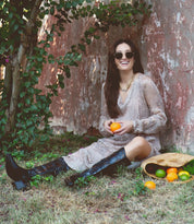 A woman with long hair and sunglasses sits on the ground outdoors, holding an orange. She is wearing a patterned dress and Bed Stu's Meadow cowgirl boots, featuring intricate floral inlay designs. A basket with citrus fruits rests beside her.