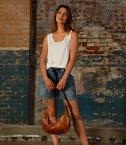 A woman stands against a weathered wall, wearing a white tank top, denim shorts, and holding the Bed Stu Megara brown shoulder bag with elegant leather details.