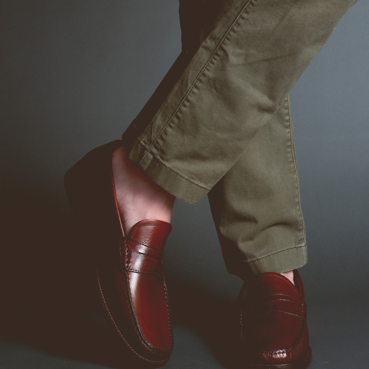 A person wearing brown Bed Stu Moat loafers and green pants, with legs crossed, stands against a dark background, exuding timeless sophistication.