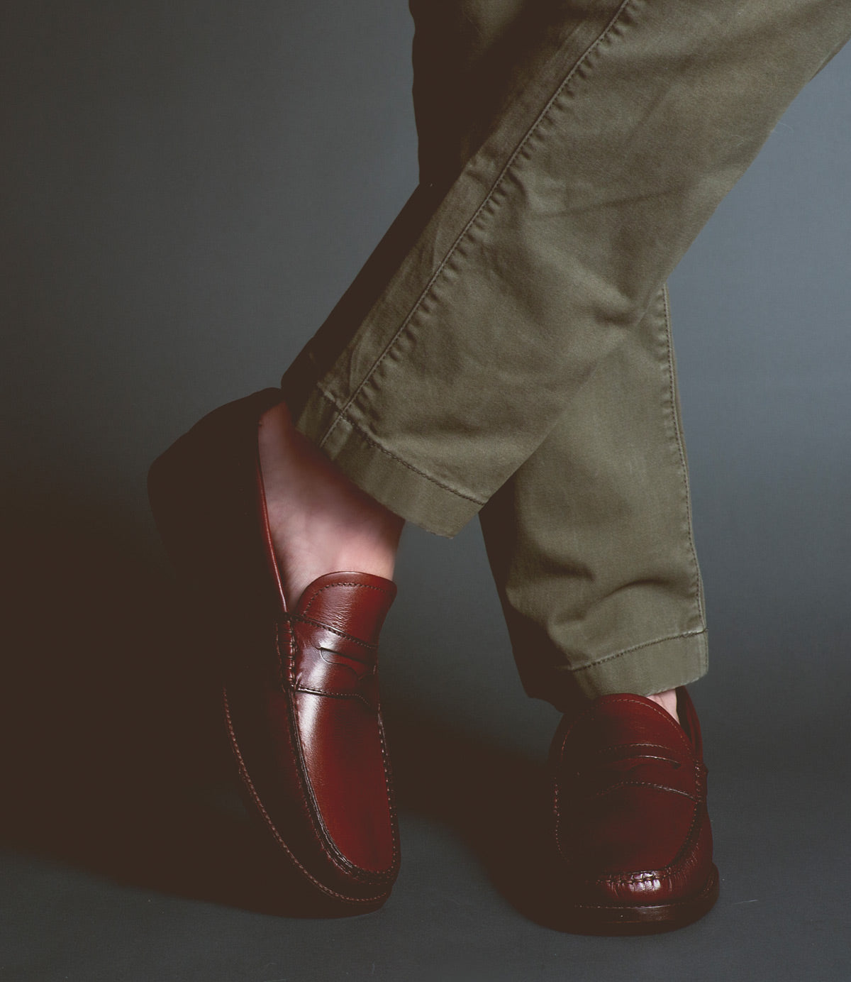 A person wearing brown Bed Stu Moat loafers and green pants, with legs crossed, stands against a dark background, exuding timeless sophistication.