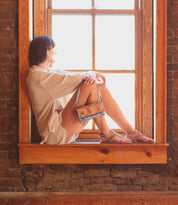 A woman sitting on a window sill, her strappy sandals adorned with ankle buckles catching the light of the moon, holds a Bed Stu purse by her side.