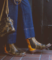 Person wearing brown Nandi Chelsea boots from Bed Stu, blue jeans, and carrying a worn leather handbag.