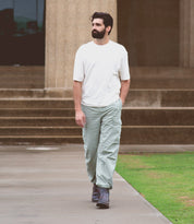 A man with a beard, wearing a white shirt and light green pants, walks on a paved path near stone steps and a grassy area. His look is completed with stylish Nando boots by Bed Stu featuring a Vibram® outsole, perfect for both comfort and durability.