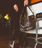 A person in a black outfit holding a Packet LTC leather handbag by Bed Stu, standing near a bar stool in a dimly lit room.