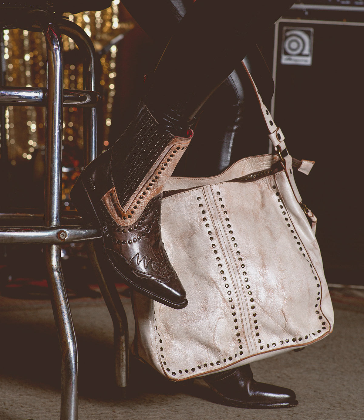 A person wearing black leather pants and brown cowboy boots sits on a stool with the Packet LTC, a large beige shoulder tote bag from Bed Stu, adorned with quality leather and metal studs resting on the floor.