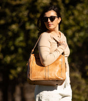 A woman wearing sunglasses carries a Bed Stu Packet LTC tan leather travel tote adorned with metal studs outdoors.