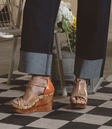 Someone wearing Bed Stu's Princess tan wedge sandals with strappy uppers and dark rolled-up jeans stands on a checkered rug. A metal container filled with flowers adds a touch of nature to this stylish scene.