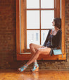 A woman sitting on a window sill with Bed Stu Princess sustainable wooden wedge shoes.