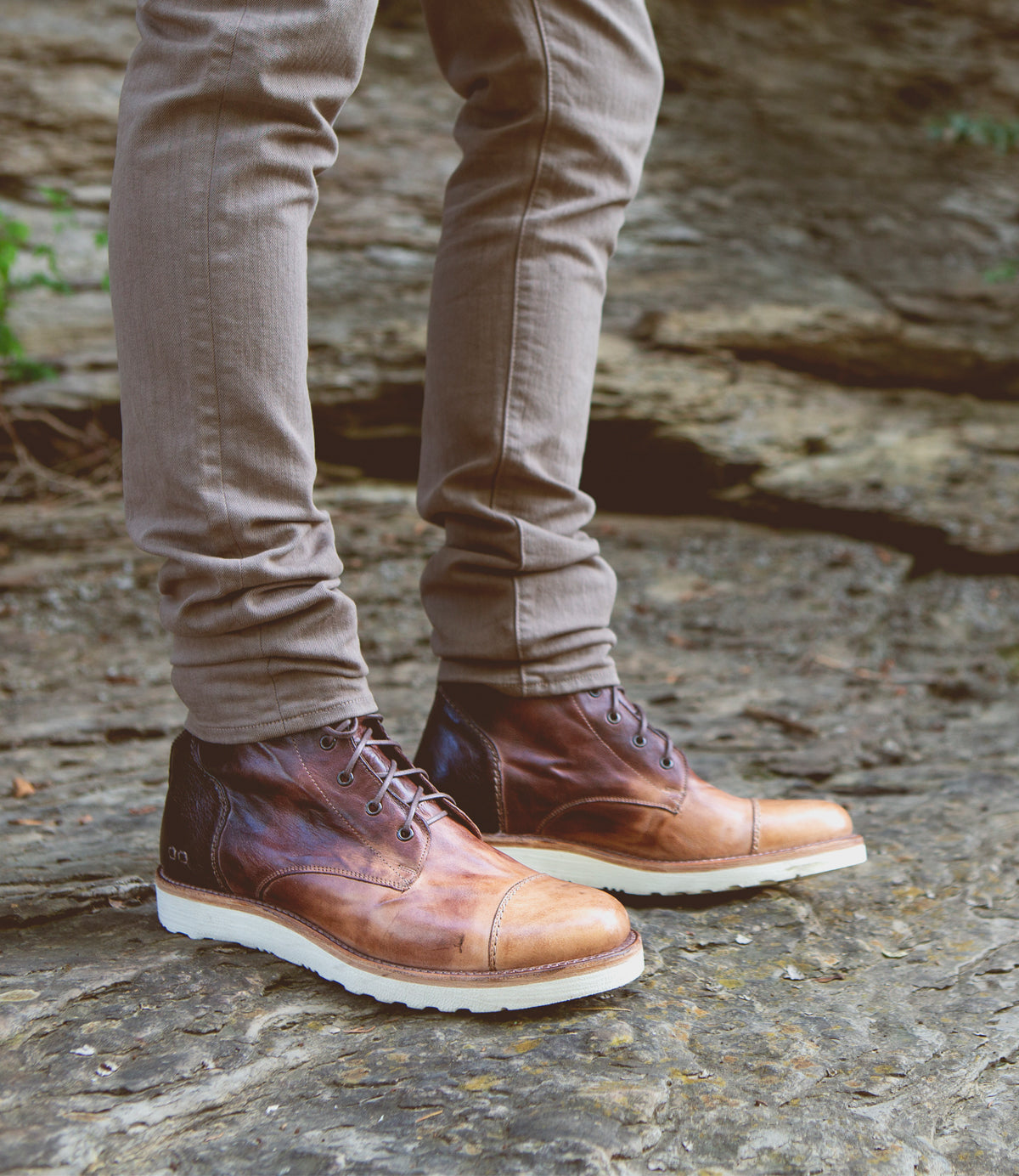 A person wearing Bed Stu men's leather boots and beige pants standing on a rocky surface.
