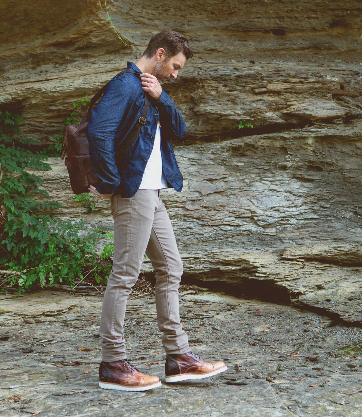 A man wearing a denim jacket, white shirt, beige pants, and Bed Stu men's leather boots stands thoughtfully on a rocky terrain, holding a Bed Stu leather bag.