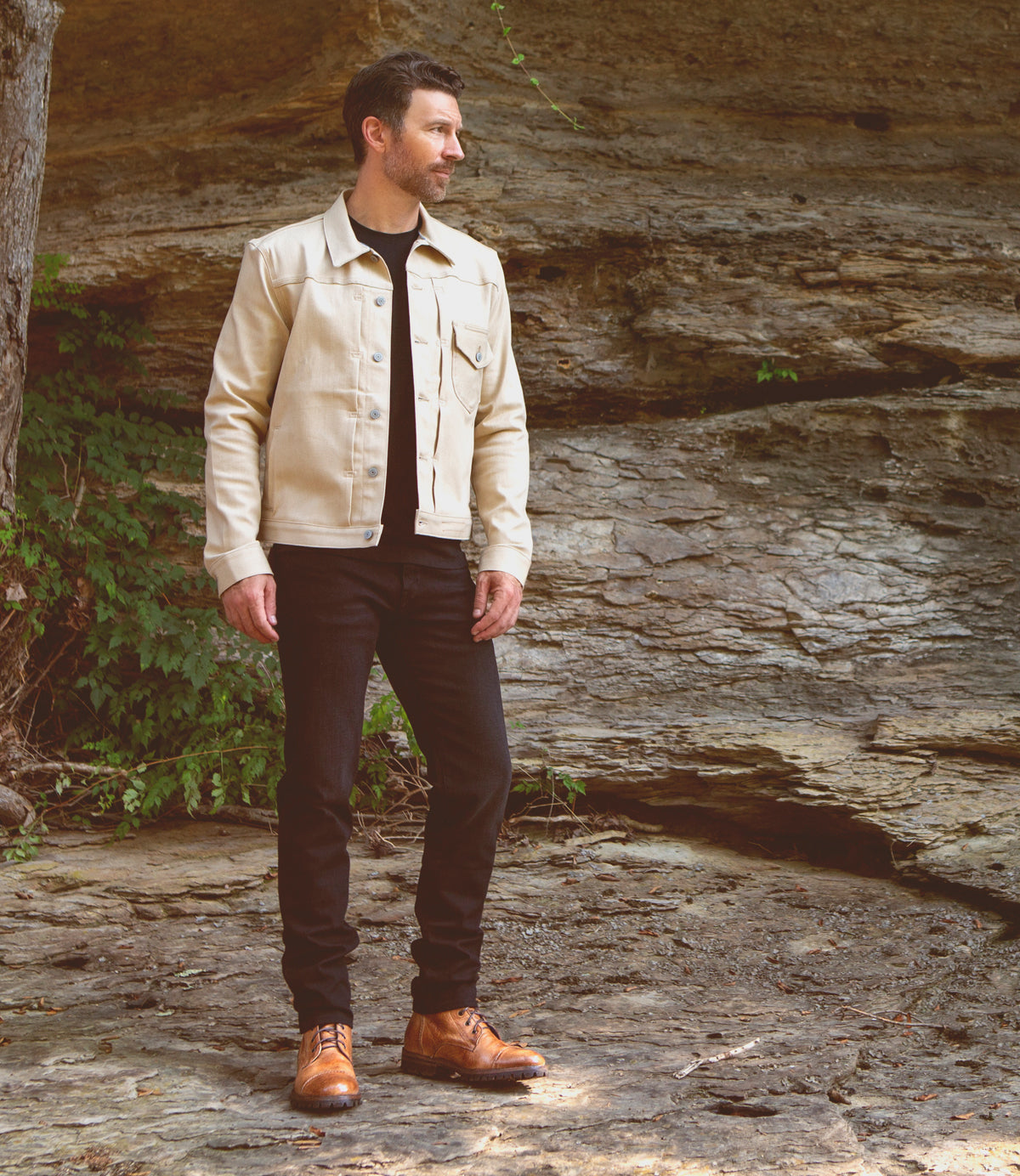 A man in a cream jacket, black jeans, and Bed Stu Protege Trek boots stands on a rocky terrain, looking to the side.