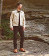 A man in a cream jacket, black jeans, and Bed Stu Protege Trek boots stands on a rocky terrain, looking to the side.