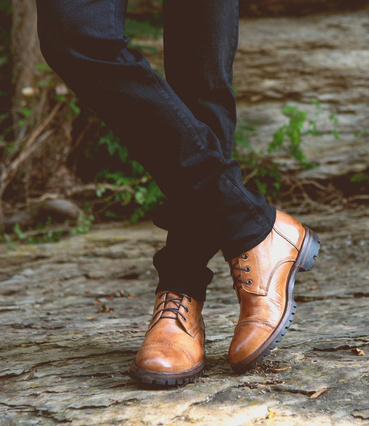 Close-up of a person's legs walking, wearing Bed Stu Protege Trek Teak Black Rustic Rust BFS Boots on a cobblestone path.