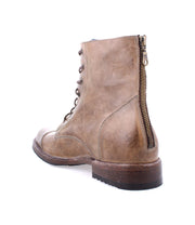 The Bed Stu Protege is a brown leather combat boot featuring a rear zipper, lace-up front, wooden sole, and Goodyear welt construction for durability. It offers robust traction plate benefits and is viewed from the back-left angle against a white background.