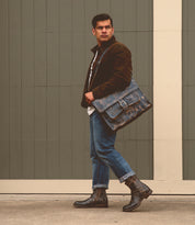 A man in a brown jacket, white shirt, and jeans stands holding the Barton by Bed Stu in front of a gray wooden wall, sporting leather high-top sneakers with a breathable insole.