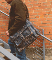 Wearing a brown jacket and leather high-top Bed Stu Barton sneakers, a person strides up the stairs beside a brick wall while carrying a weathered leather satchel.
