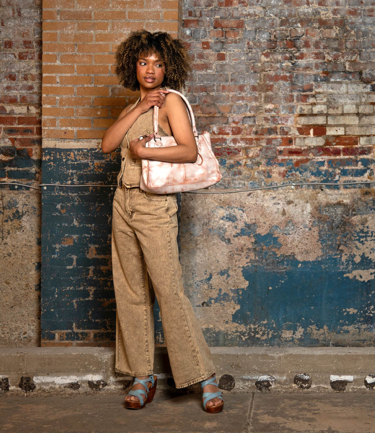 A woman stands against a distressed brick wall, wearing tan overalls and platform sandals, holding the Bed Stu Rockababy compact bag with its versatile crossbody strap.