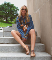 A woman wearing sunglasses, a blue shirt, and buttery soft leather brown Rumba II shoes by Bed Stu sits on outdoor steps, smiling. There are trees and cars in the background.