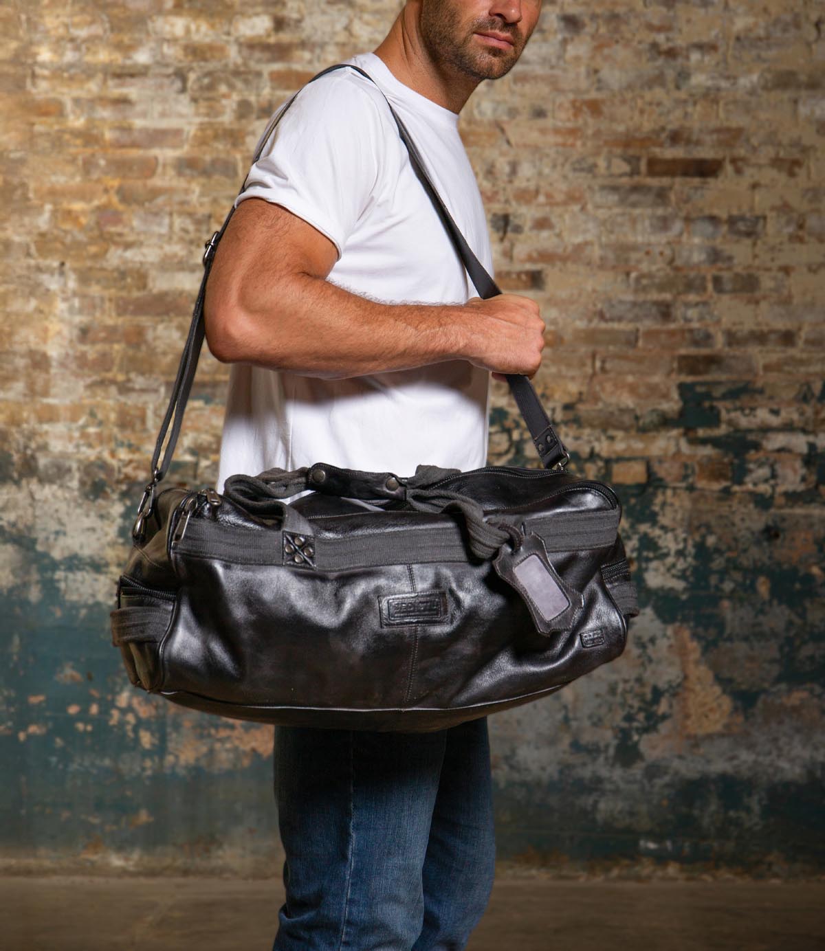 A man in a white t-shirt and jeans stands against a worn brick wall, carrying the Bed Stu Ruslan, a large black leather duffle, over his shoulder.