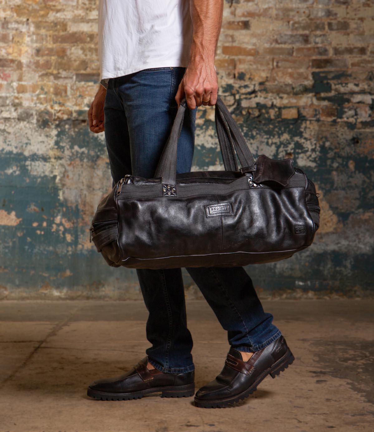 A person in jeans and loafers stands indoors against a distressed brick wall, holding the Bed Stu Ruslan black leather duffle travel bag by its handles.
