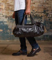 A person in jeans and loafers stands indoors against a distressed brick wall, holding the Bed Stu Ruslan black leather duffle travel bag by its handles.