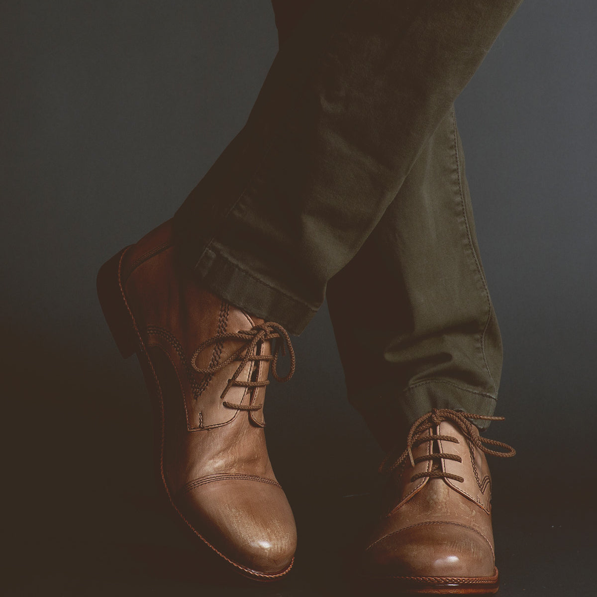 A person wearing dark green pants and Scoot rugged lace-up boots by Bed Stu, handcrafted in Italy, stands with legs crossed against a dark background.