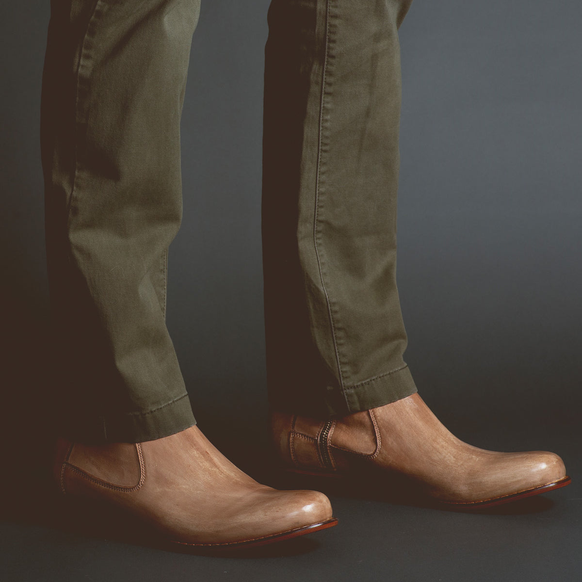 A person wearing olive green pants and light brown vegetable-tanned Bed Stu Seek slip-on shoes, showcasing artisanal craftsmanship, stands against a dark background.