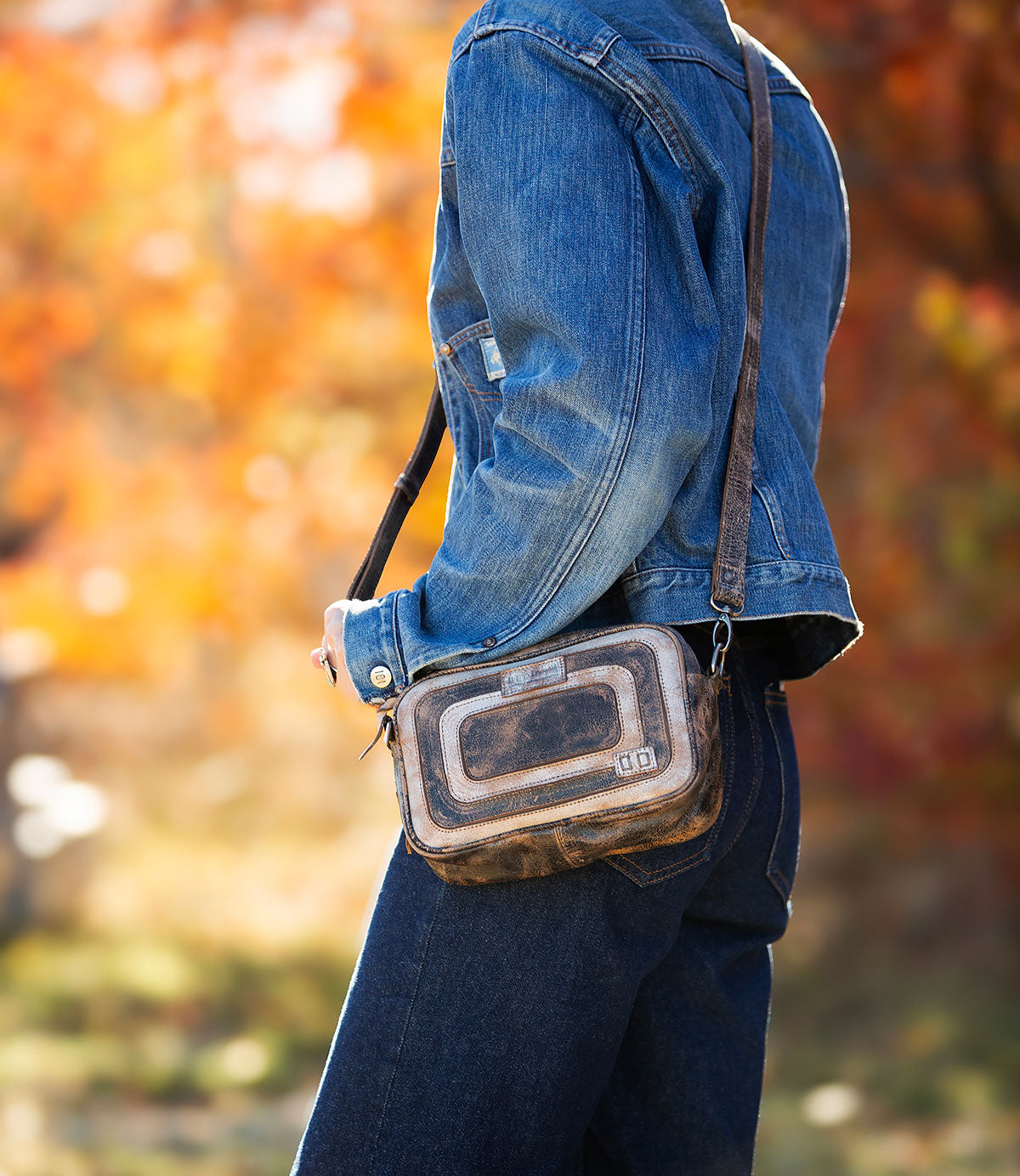 A person in a denim jacket and jeans strolls through autumn foliage, carrying the Bed Stu "Shy," a compact crossbody bag featuring contrasting leather details.