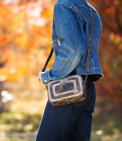 A person in a denim jacket and jeans strolls through autumn foliage, carrying the Bed Stu "Shy," a compact crossbody bag featuring contrasting leather details.