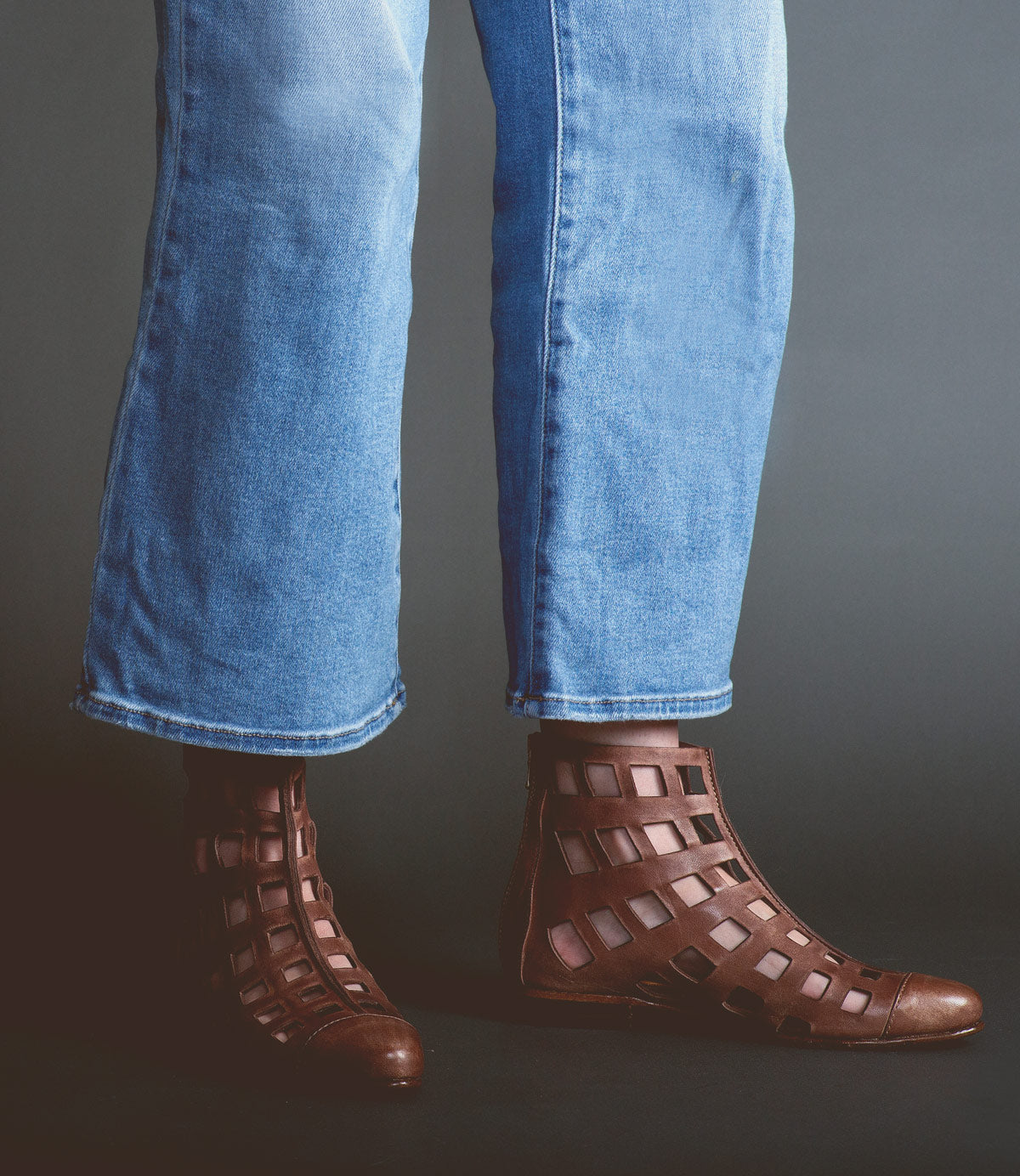 Person wearing blue jeans and edgy and chic brown lattice-pattern leather ankle boots, Skyline by Bed Stu, handcrafted in Italy, standing against a dark background.