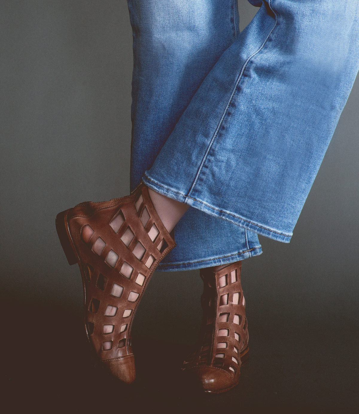 Person wearing flare jeans and **Skyline** brown ankle boots handcrafted in Italy by **Bed Stu**, featuring bold square cutouts, standing with one leg slightly crossed over the other.