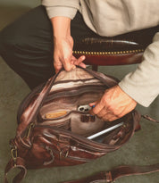 Sitting on a chair, someone reaches for their Bed Stu Barton brown leather bag, placing a phone, notebook, and other essentials inside. Nearby, sleek leather high-top sneakers rest on the floor, ready to be worn.