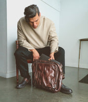 Seated in a beige sweater and black pants, the person peers into a brown leather bag with a laptop partially visible. The look is completed with stylish Barton men's leather high-top sneakers by Bed Stu, featuring breathable insoles for comfort and flair.