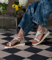 A person in floral jeans and beige Soni II platform sandals by Bed Stu, featuring an XL EXTRALIGHT® outsole, stands on a checkered rug with flowers in a metal box behind them.