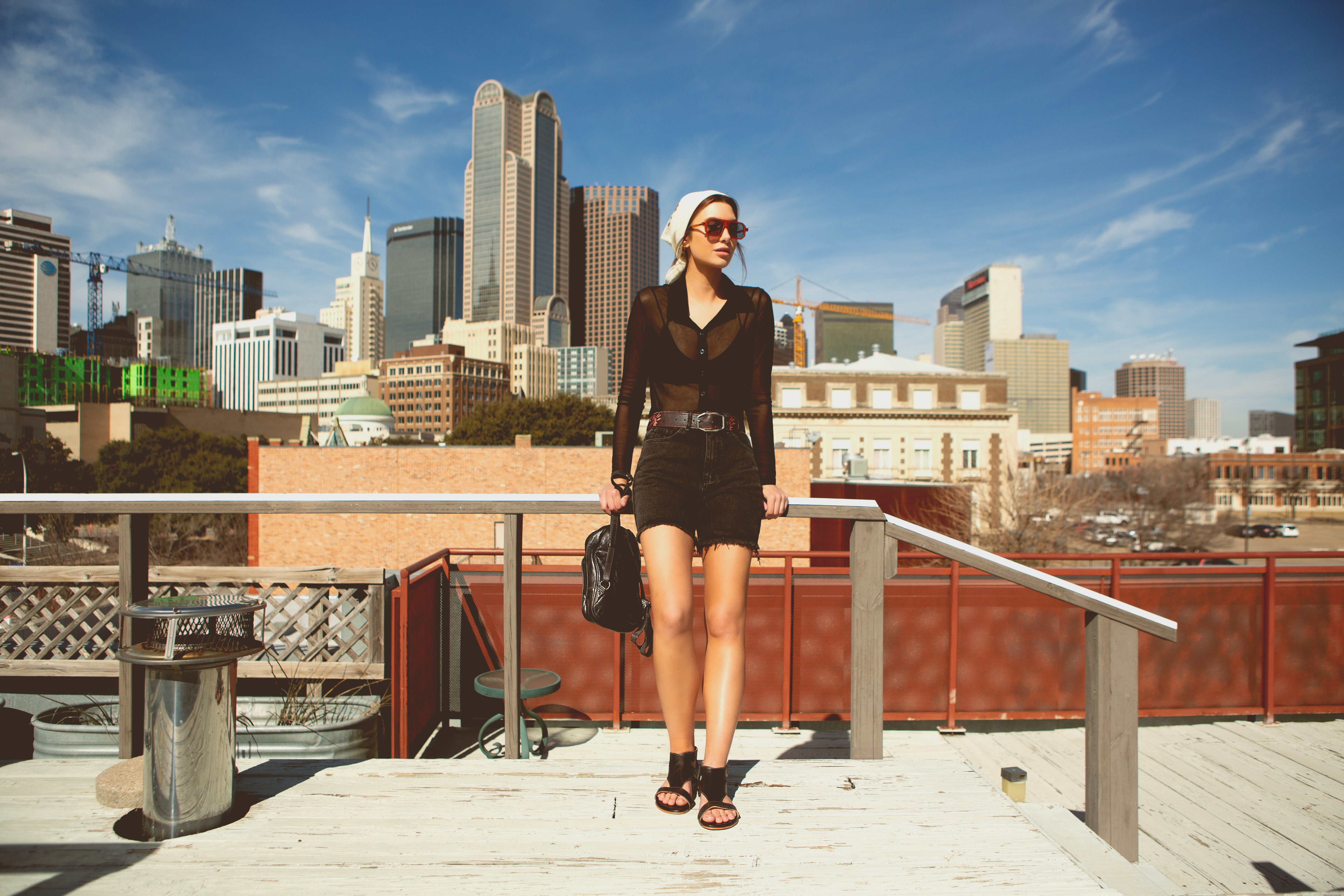 A woman in casual attire stands on a rooftop with a city skyline in the background on a clear day. Sporting trendy accessories, she wears sunglasses, a black top, and shorts while holding a black bag. Complete Your Look with Our 'Shop the Look' Bundle! by Bed|Stü for this effortlessly chic ensemble.