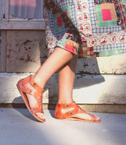 Woman wearing Bed Stu Soto Cutout flat leather sandals and patterned dress standing against a weathered wall.