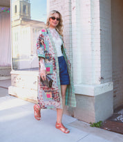 A woman in a colorful patchwork coat and sunglasses, posing on a city sidewalk, wearing Bed Stu Soto Cutout leather sandals.