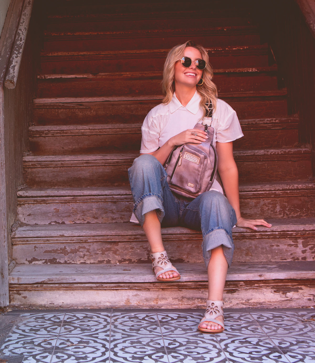 A woman in sunglasses sits smiling on a staircase, holding a backpack and wearing Bed Stu vegetable tanned leather Soto Cutout flat leather sandals.