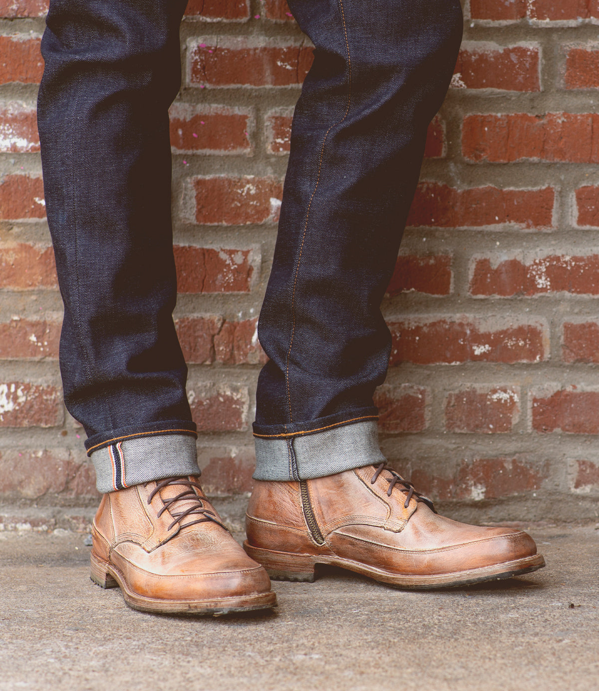A person wearing Bed Stu Spiker Teak Rustic Boots and cuffed jeans standing against a brick wall.