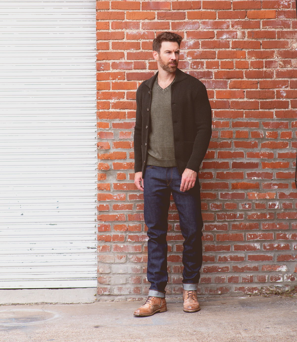 A man standing against a brick wall, wearing a dark cardigan, green shirt, blue jeans, and brown leather Bed Stu lace-up ankle boots.