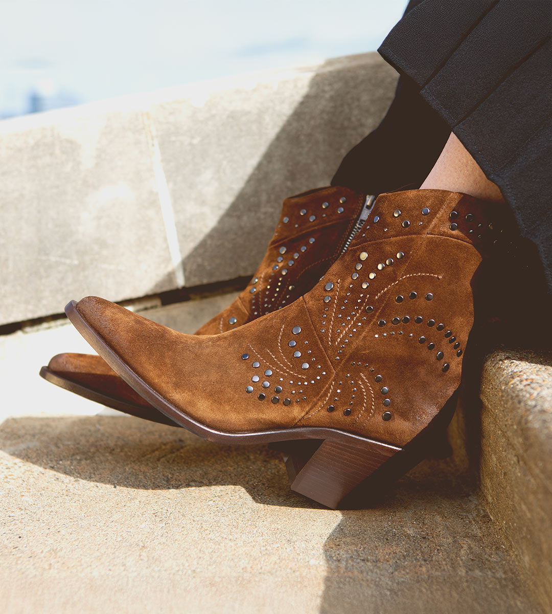 The person sitting on stone steps is wearing the Sposa by Bed Stu, featuring brown suede ankle boots with a leg-lengthening effect, decorative stitching, and studded details.