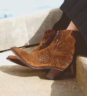 The person sitting on stone steps is wearing the Sposa by Bed Stu, featuring brown suede ankle boots with a leg-lengthening effect, decorative stitching, and studded details.