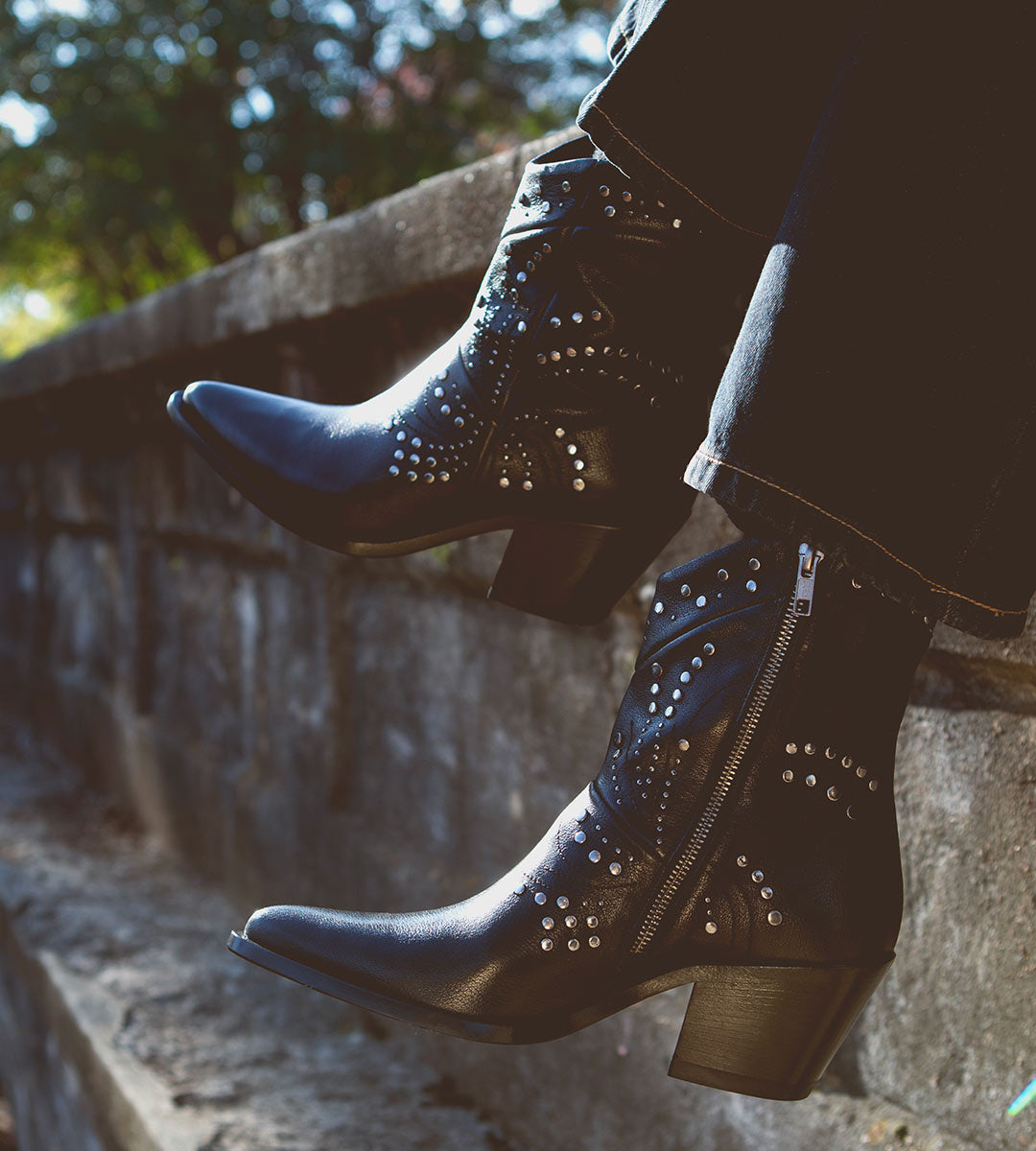 A person wearing Sposa booties by Bed Stu, crafted from black Italian leather with studded details and pointed toes, sits on a stone ledge.