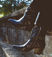 A person wearing Sposa booties by Bed Stu, crafted from black Italian leather with studded details and pointed toes, sits on a stone ledge.